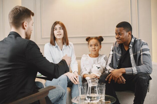 Couple with a young girl holding a teddy bear talking to an adviser on a sofa