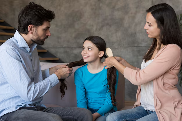 Parents brushing and holding a smiling girl's hair while sitting on a sofa