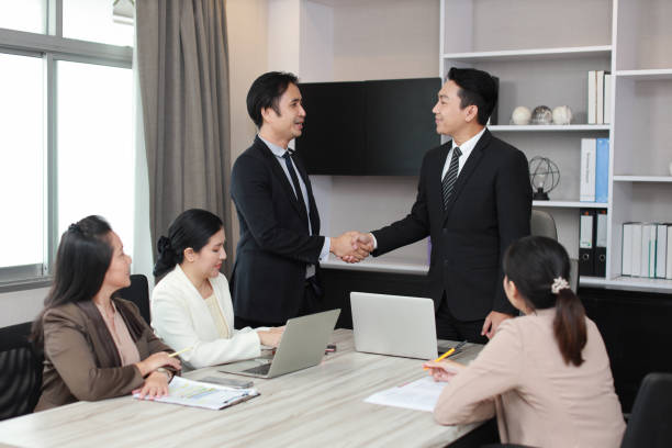 Two businessmen shaking hands in a meeting room while colleagues watch