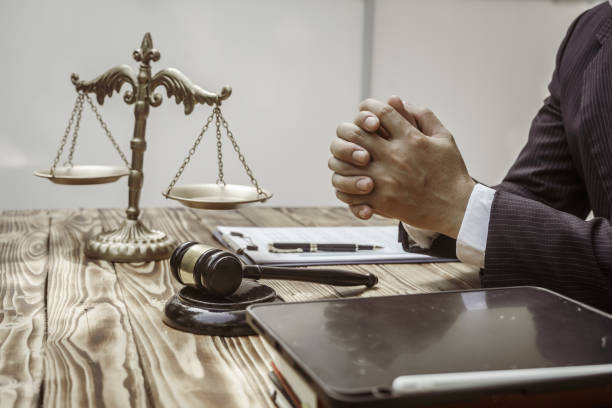 Man in a suit with clasped hands at a desk beside a gavel and scales of justice