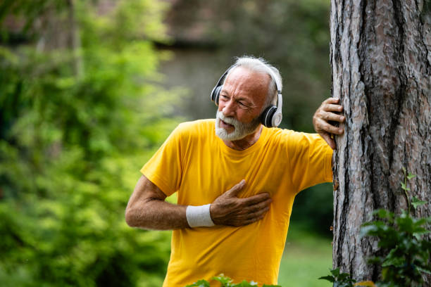 Older man in headphones leaning on a tree with a hand pressed to his chest