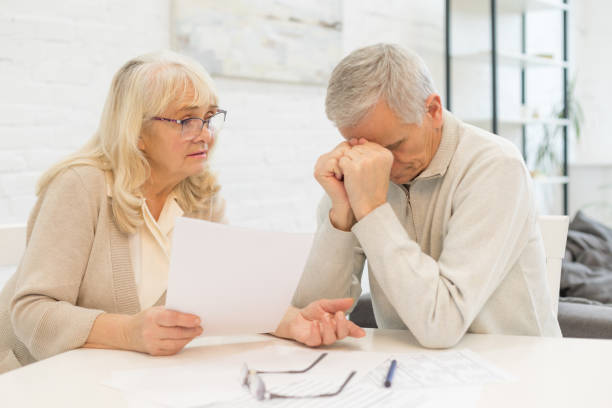 Older woman holding a document beside a distressed older man at a table