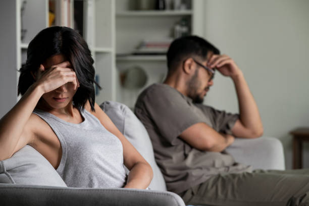 Couple sitting apart on a sofa, each holding their head in frustration