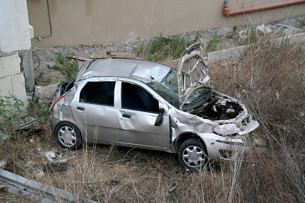 Crumpled silver hatchback with its hood up in weeds below a retaining wall