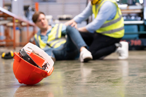 Orange hard hat on a warehouse floor with an injured worker in a safety vest behind