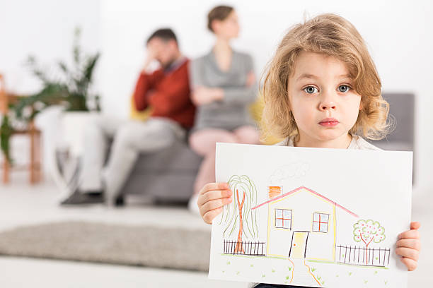 Young child holding a drawing of a house while parents sit apart behind
