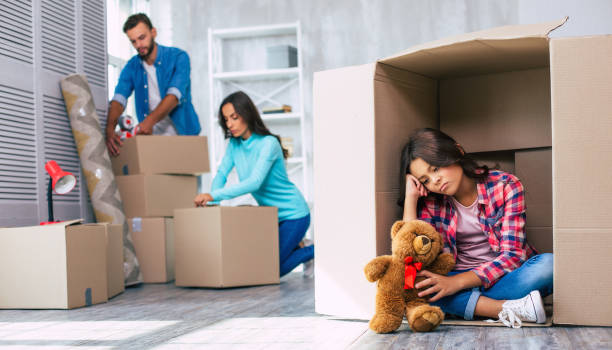 Sad girl holding a teddy bear inside a moving box while parents pack